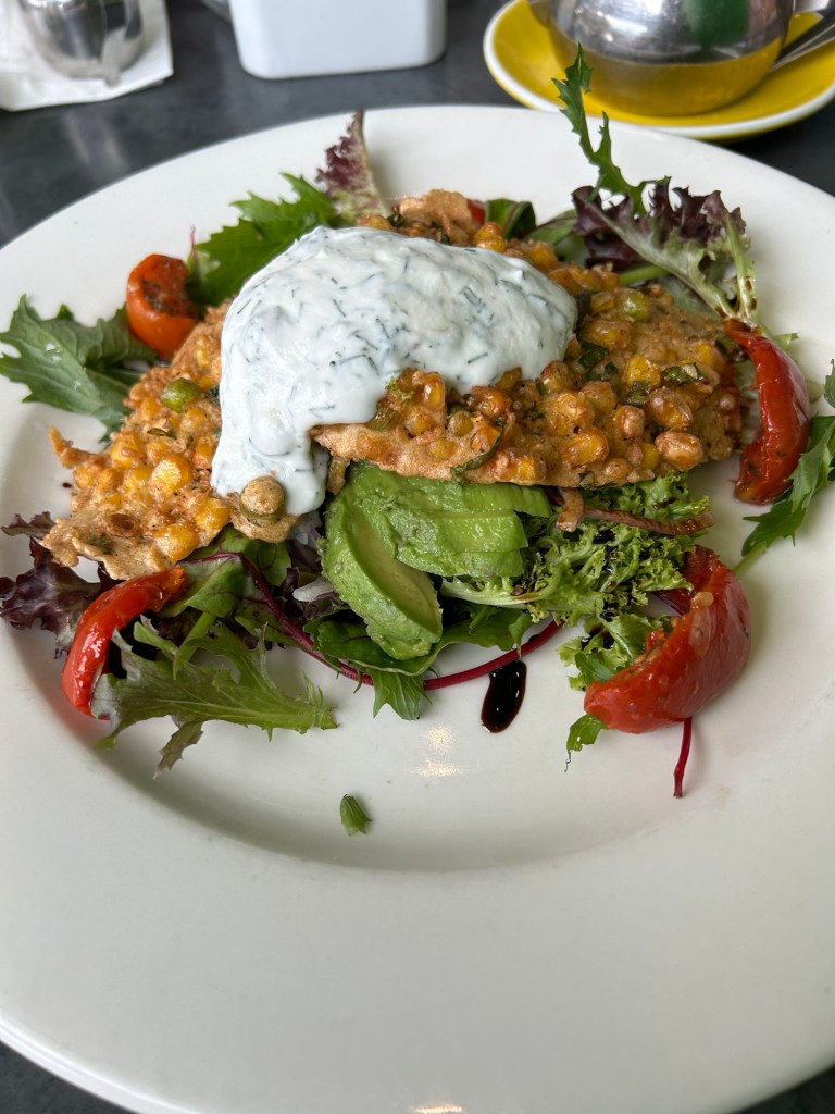 A plated dish featuring a fried protein patty topped with a creamy sauce, surrounded by a colorful salad of mixed greens, sliced avocado, and cherry tomatoes.