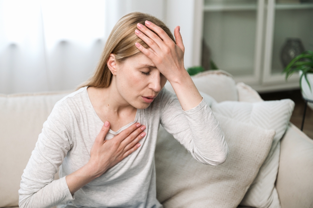 A woman sitting on a couch, appearing distressed, with her hand on her forehead and another hand on her chest, indicating discomfort or anxiety.