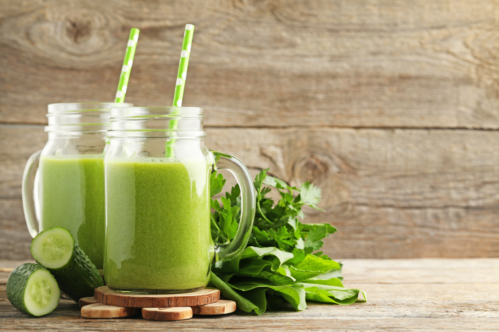 Two jars of green smoothie with striped straws, surrounded by fresh cucumber and leafy greens on a wooden surface.