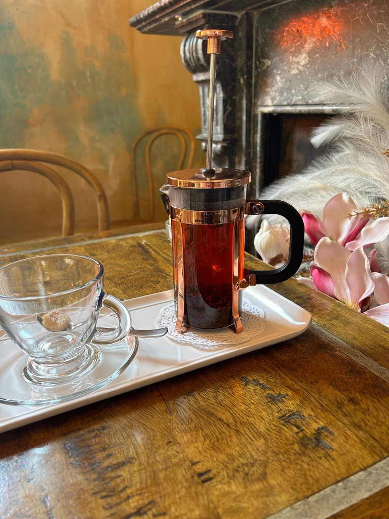 A French press with brewed tea sits on a tray beside an empty glass cup and a piece of sugar, with flowers and a rustic wooden table in the background.