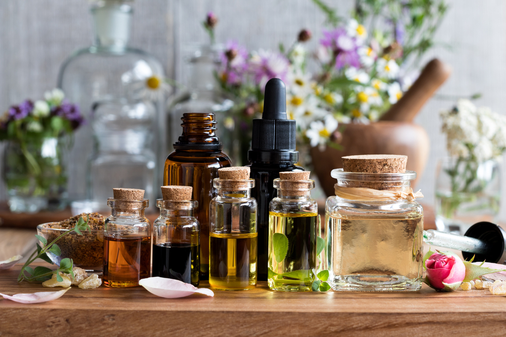 A wooden table displaying an assortment of glass bottles filled with various essential oils, next to jars with natural ingredients, surrounded by fresh flowers and herbs.