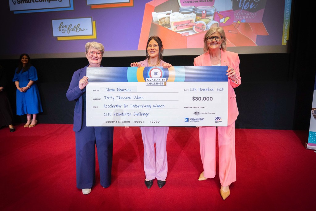 Three women standing on stage holding a large cheque for $30,000, awarded for the 2023 Kickstarter Challenge, with a backdrop displaying event branding.