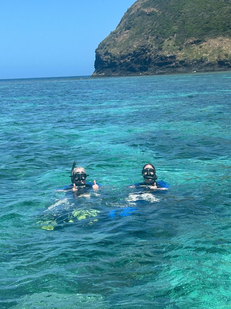 Two individuals snorkeling in clear blue water with a rocky coastline in the background.