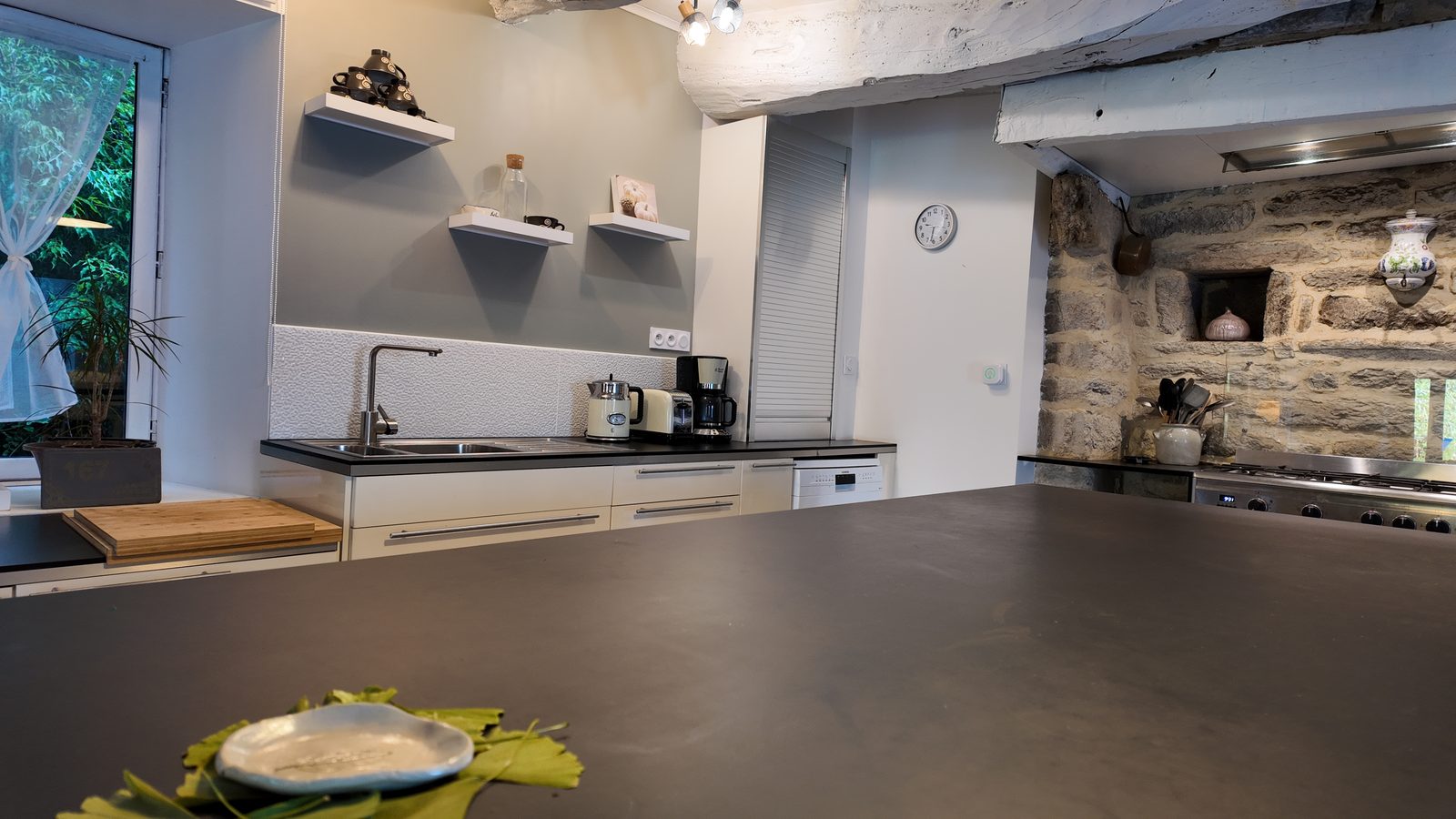 La cuisine vue depuis l’îlot, l’évier à gauche et le piano de cuisson devant la cheminée de pierre