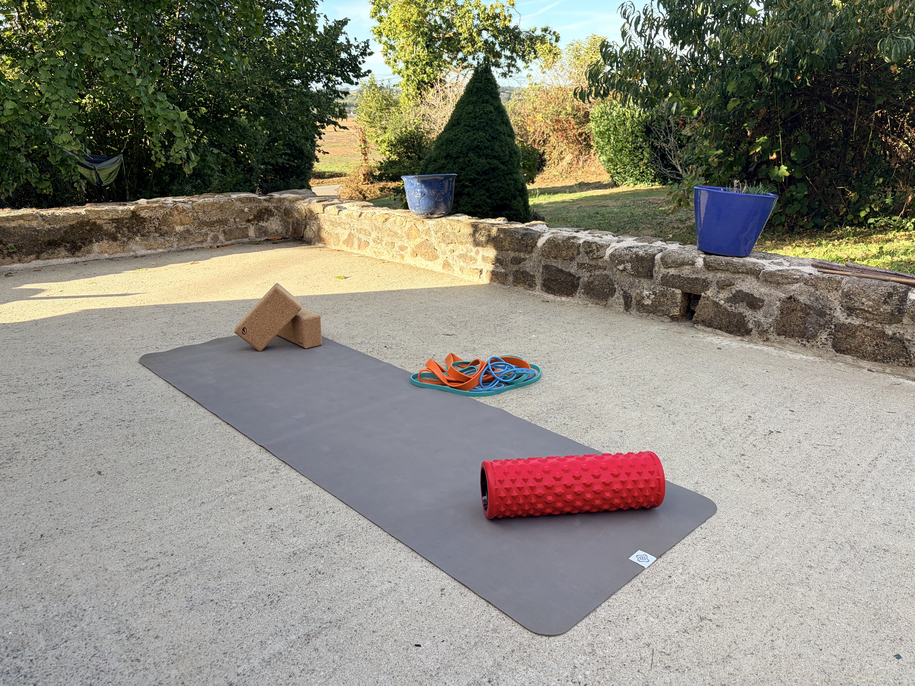 Un tapis de yoga déroulé sur la terrasse, briques et rouleau posés à côté, le jardin derrière